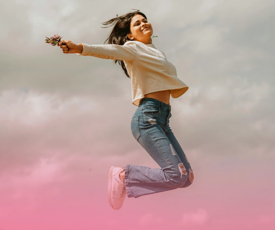 Woman joyfully jumping with flowers under cloudy sky.