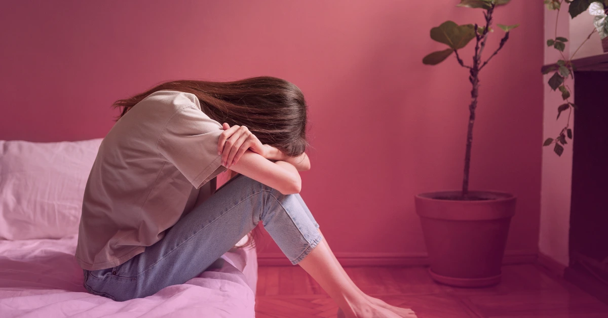 Woman sitting on bed, appearing distressed, near plant.